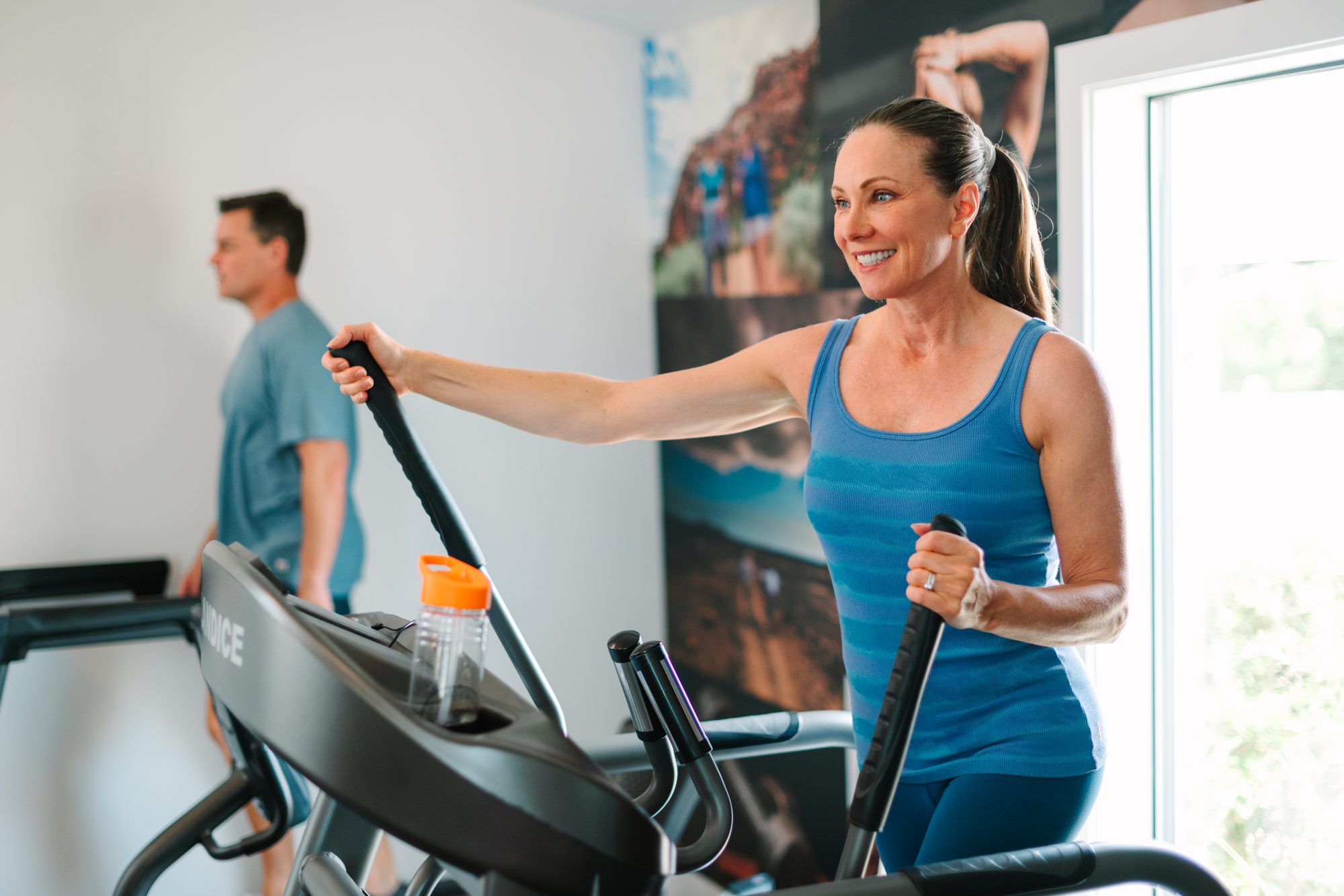 A smiling woman in a blue tank top uses an elliptical machine in a gym, while a man exercises in the background in Phoenix